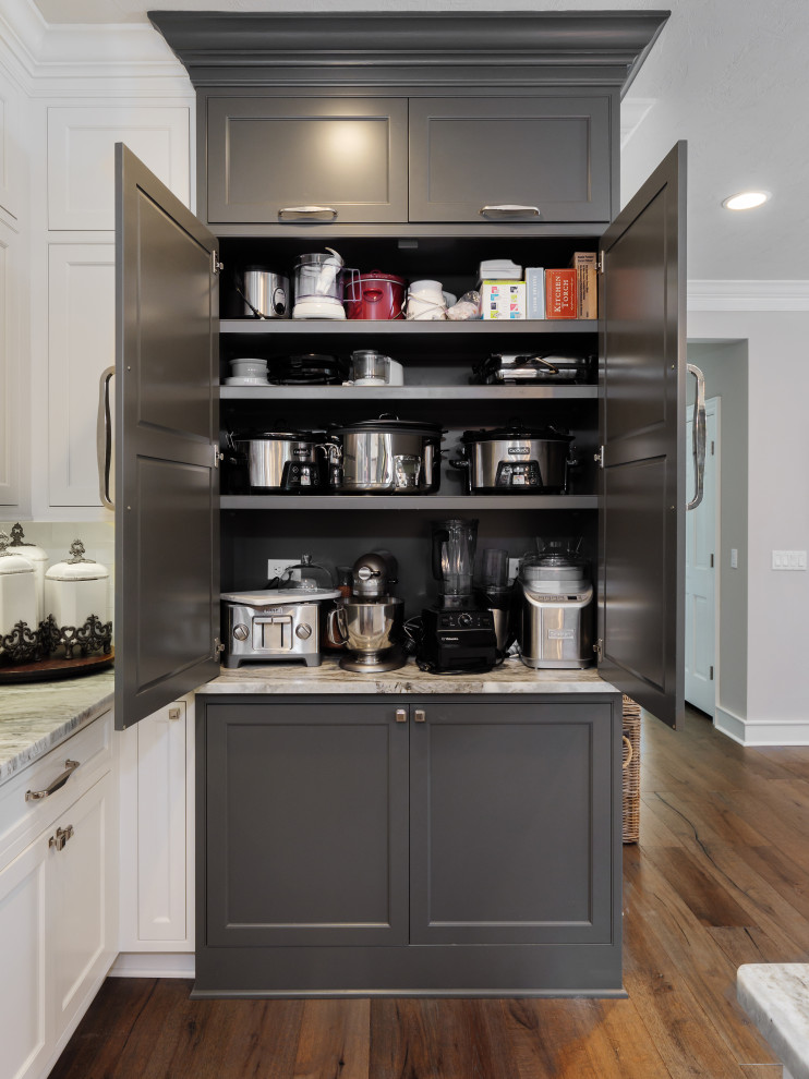 Klassisk inredning av ett stort grå grått kök, med en rustik diskho, luckor med infälld panel, vita skåp, bänkskiva i kvartsit, vitt stänkskydd, stänkskydd i porslinskakel, rostfria vitvaror, mörkt trägolv, en köksö och brunt golv