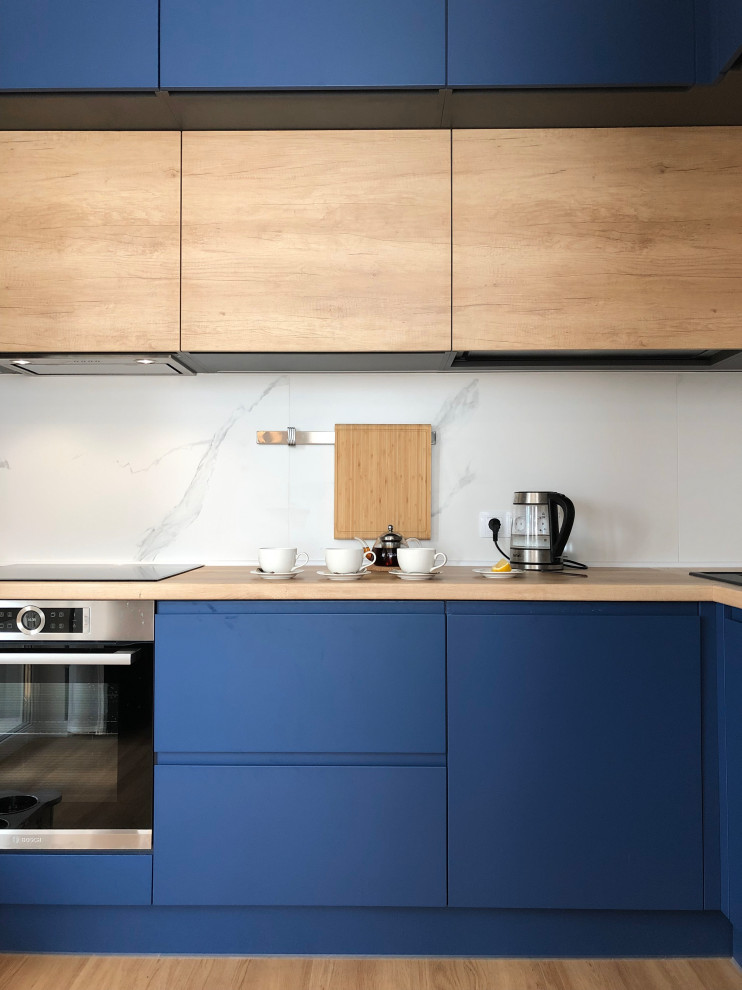 This is an example of a small scandinavian l-shaped open plan kitchen with a drop-in sink, flat-panel cabinets, blue cabinets, wood benchtops, white splashback, porcelain splashback, black appliances, vinyl floors, no island, beige floor and beige benchtop.