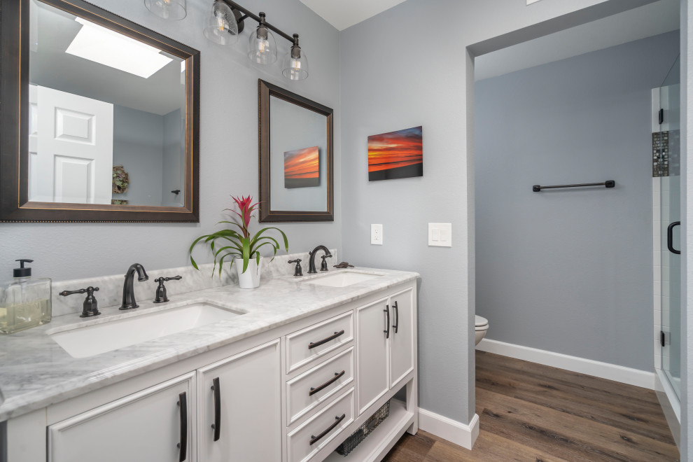 This is an example of a mid-sized transitional master bathroom in Los Angeles with furniture-like cabinets, white cabinets, a freestanding tub, an alcove shower, a two-piece toilet, multi-coloured tile, white walls, vinyl floors, an undermount sink, marble benchtops, brown floor, a hinged shower door, grey benchtops, a double vanity and a freestanding vanity.