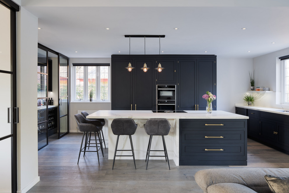 This is an example of a transitional u-shaped open plan kitchen in London with an undermount sink, beaded inset cabinets, black cabinets, black appliances, dark hardwood floors, with island, brown floor and white benchtop.