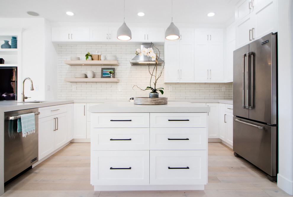Beach style u-shaped kitchen in Orange County with an undermount sink, shaker cabinets, white cabinets, white splashback, stainless steel appliances, light hardwood floors, with island, beige floor and grey benchtop.