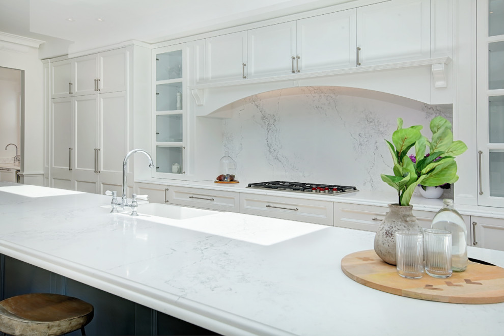 This is an example of a large contemporary galley kitchen pantry in Sydney with a farmhouse sink, shaker cabinets, white cabinets, quartz benchtops, white splashback, engineered quartz splashback, stainless steel appliances, light hardwood floors, with island, brown floor and white benchtop.