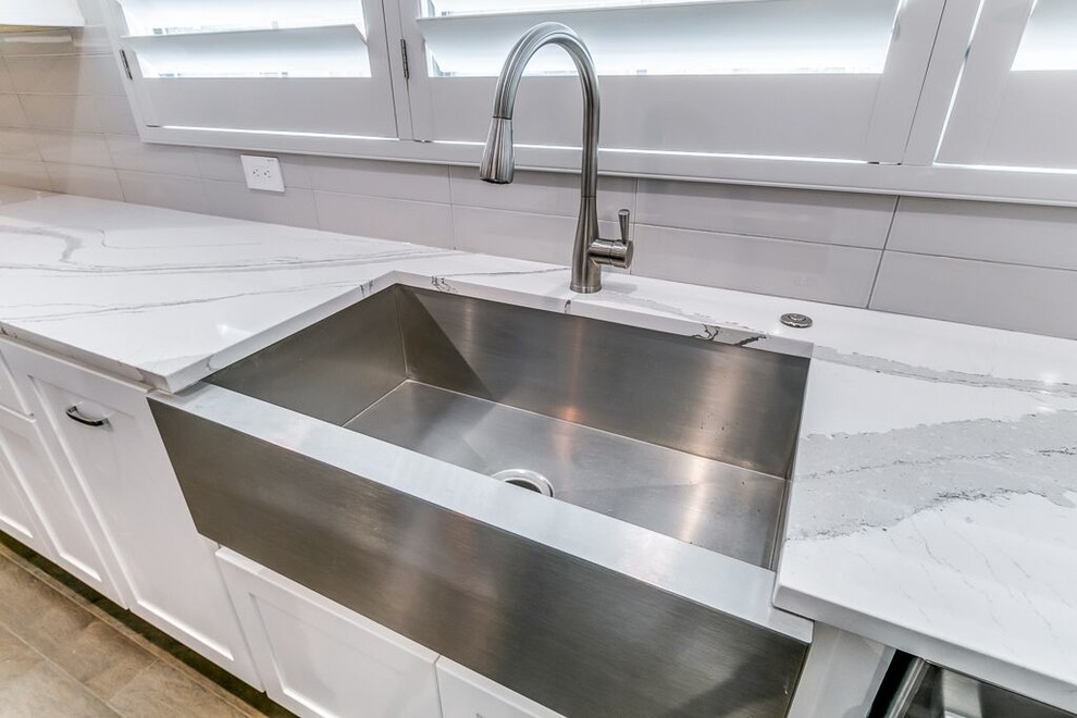 Inspiration for a transitional l-shaped brown floor open concept kitchen remodel in Houston with a farmhouse sink, shaker cabinets, white cabinets, quartz countertops, beige backsplash, subway tile backsplash, stainless steel appliances and an island