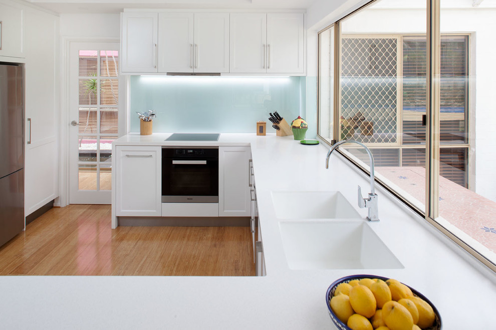 Inspiration for a mid-sized modern u-shaped kitchen pantry in Perth with an integrated sink, recessed-panel cabinets, white cabinets, solid surface benchtops, white splashback, glass sheet splashback, stainless steel appliances, bamboo floors and a peninsula.