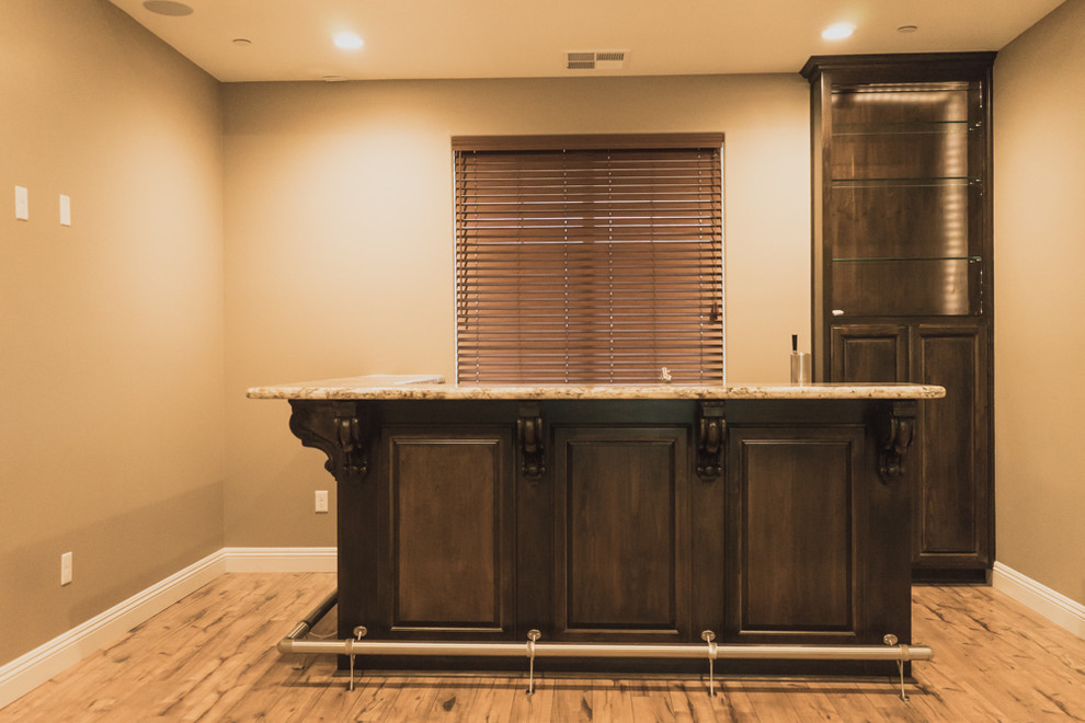 This is an example of a mid-sized traditional single-wall wet bar in Other with raised-panel cabinets, dark wood cabinets, granite benchtops, light hardwood floors and beige floor.