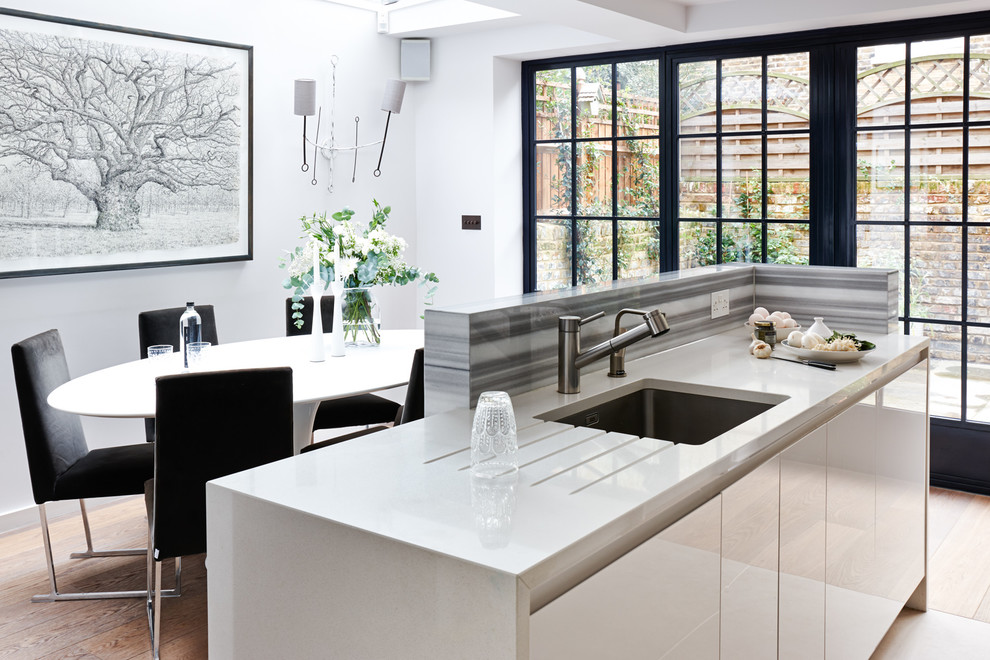This is an example of a small modern galley eat-in kitchen in London with an integrated sink, flat-panel cabinets, white cabinets, solid surface benchtops, grey splashback, glass sheet splashback, stainless steel appliances, medium hardwood floors, with island and white benchtop.