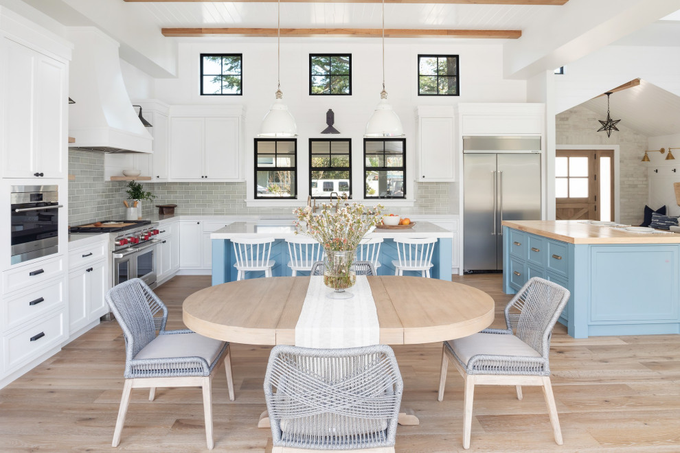 Bild på ett stort lantligt vit vitt kök, med en undermonterad diskho, skåp i shakerstil, blå skåp, bänkskiva i kvartsit, grönt stänkskydd, stänkskydd i tunnelbanekakel, rostfria vitvaror, ljust trägolv, flera köksöar och beiget golv