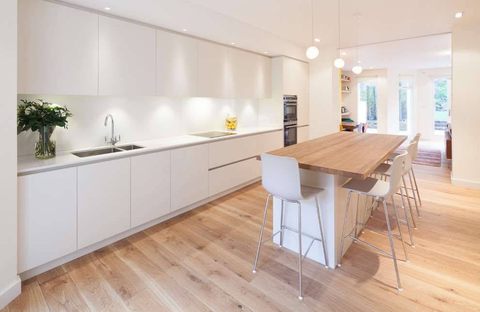 Photo of a large scandinavian single-wall open plan kitchen in London with an undermount sink, flat-panel cabinets, white cabinets, solid surface benchtops, white splashback, glass sheet splashback, with island, light hardwood floors and stainless steel appliances.