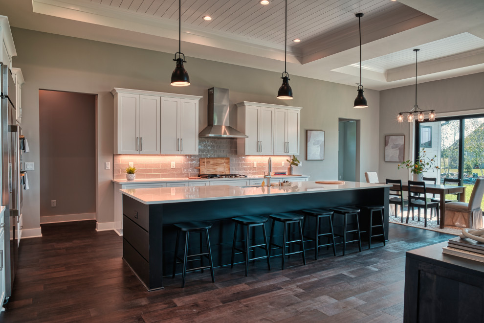 Inspiration for a large transitional l-shaped medium tone wood floor, brown floor and tray ceiling open concept kitchen remodel in Milwaukee with a farmhouse sink, shaker cabinets, white cabinets, quartz countertops, gray backsplash, subway tile backsplash, stainless steel appliances, an island and white countertops