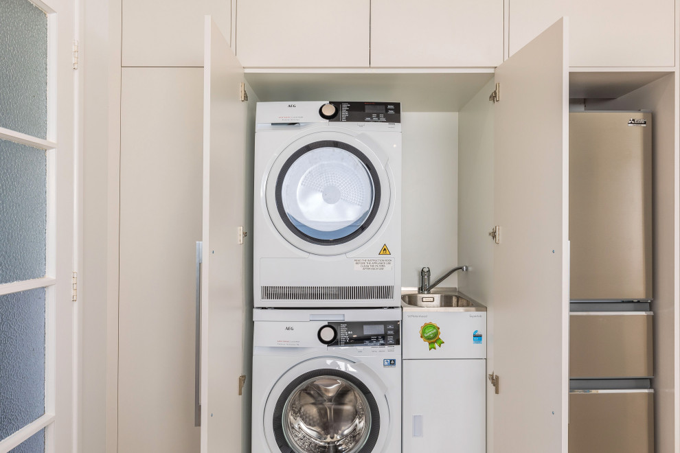 Immagine di un piccolo ripostiglio-lavanderia minimal con lavatoio, ante lisce, ante bianche, pareti bianche, pavimento in laminato, lavatrice e asciugatrice a colonna e pavimento marrone
