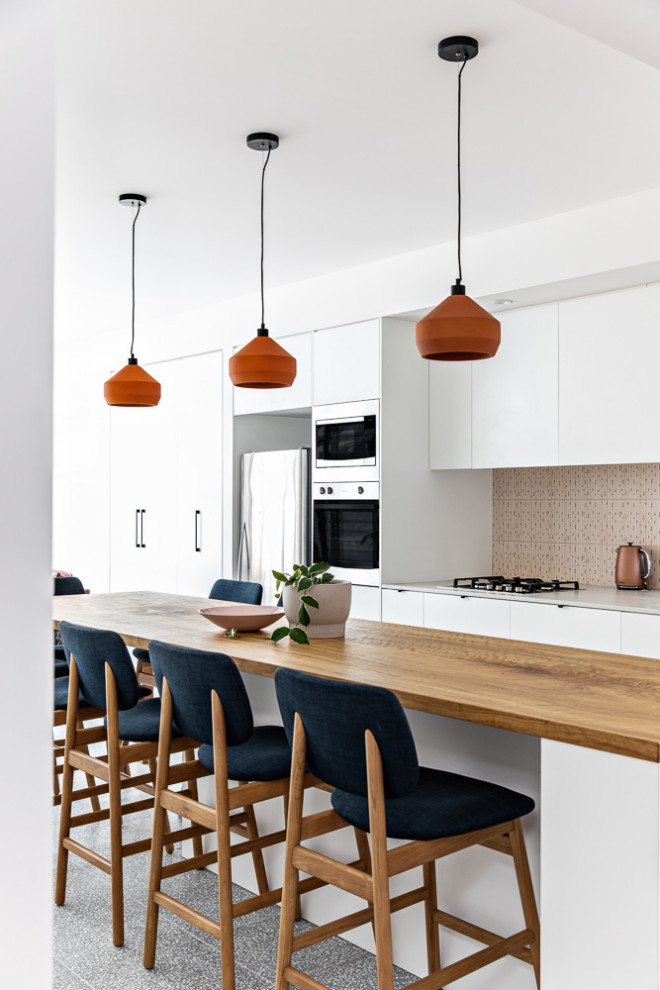 This is an example of a small contemporary galley open plan kitchen in Sydney with an undermount sink, white cabinets, wood benchtops, pink splashback, ceramic splashback, stainless steel appliances, terrazzo floors, with island, grey floor and brown benchtop.
