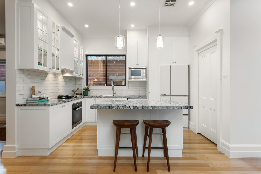 Inspiration for a contemporary l-shaped kitchen in Melbourne with white splashback, multi-coloured benchtop, shaker cabinets, white cabinets, ceramic splashback, white appliances, light hardwood floors, with island and beige floor.