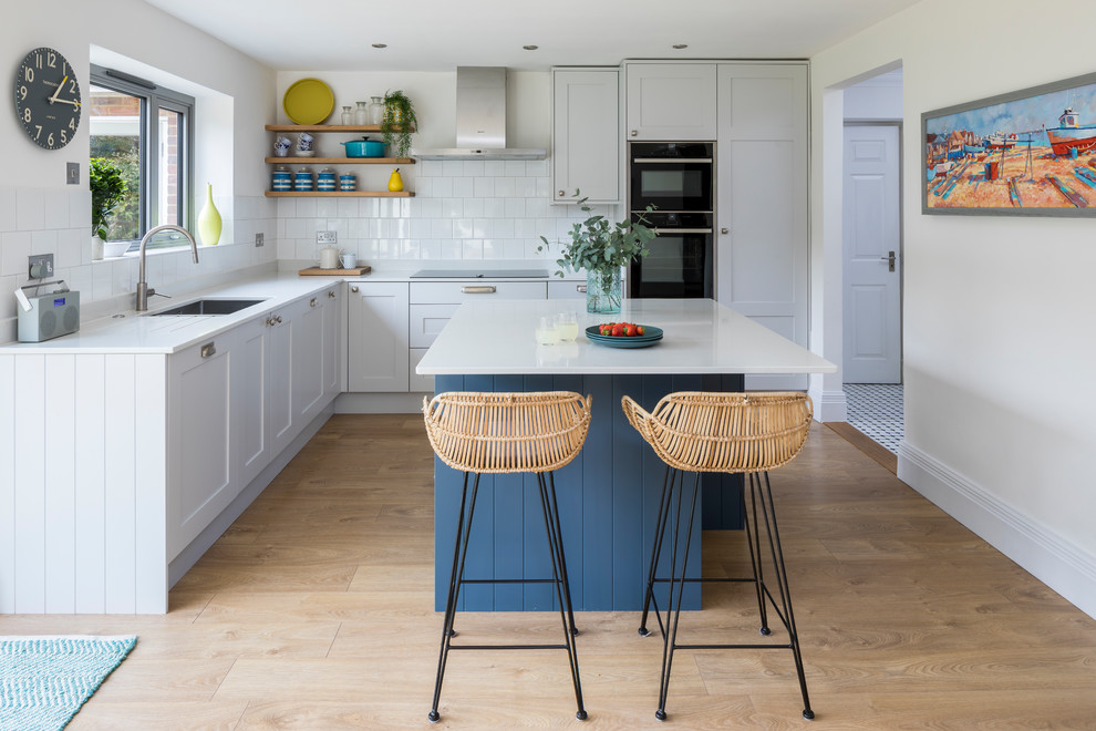 This is an example of a mid-sized contemporary l-shaped open plan kitchen in Surrey with an undermount sink, shaker cabinets, blue cabinets, solid surface benchtops, white splashback, porcelain splashback, stainless steel appliances, light hardwood floors, with island, beige floor and white benchtop.