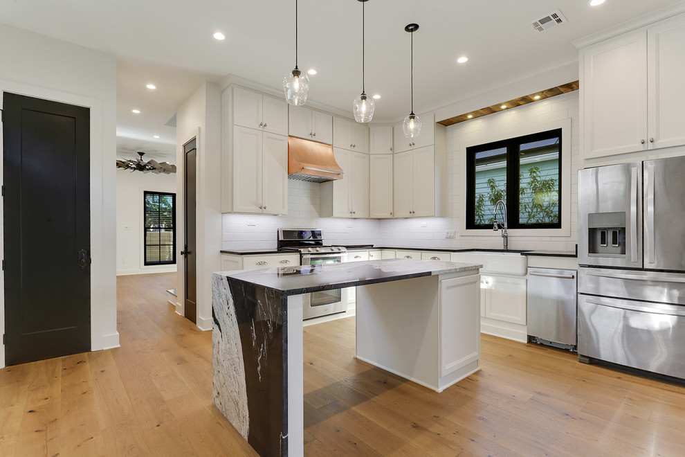 This is an example of a small country l-shaped eat-in kitchen in New Orleans with a farmhouse sink, flat-panel cabinets, white cabinets, granite benchtops, white splashback, subway tile splashback, stainless steel appliances, light hardwood floors, with island, brown floor and black benchtop.