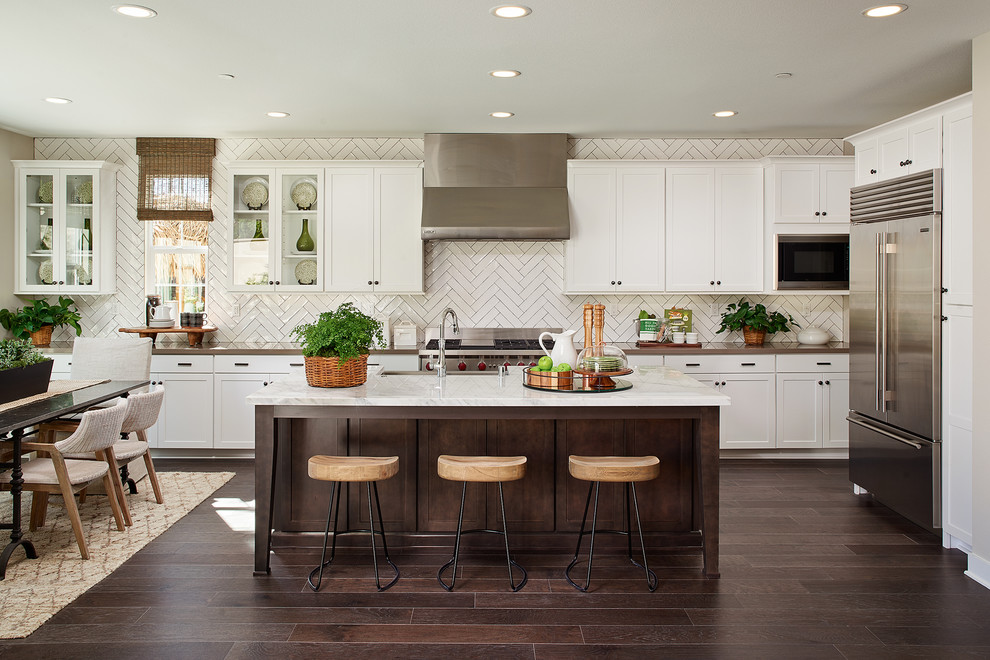 Design ideas for a large traditional galley open plan kitchen in San Diego with a farmhouse sink, glass-front cabinets, white cabinets, marble benchtops, white splashback, porcelain splashback, stainless steel appliances, dark hardwood floors and with island.