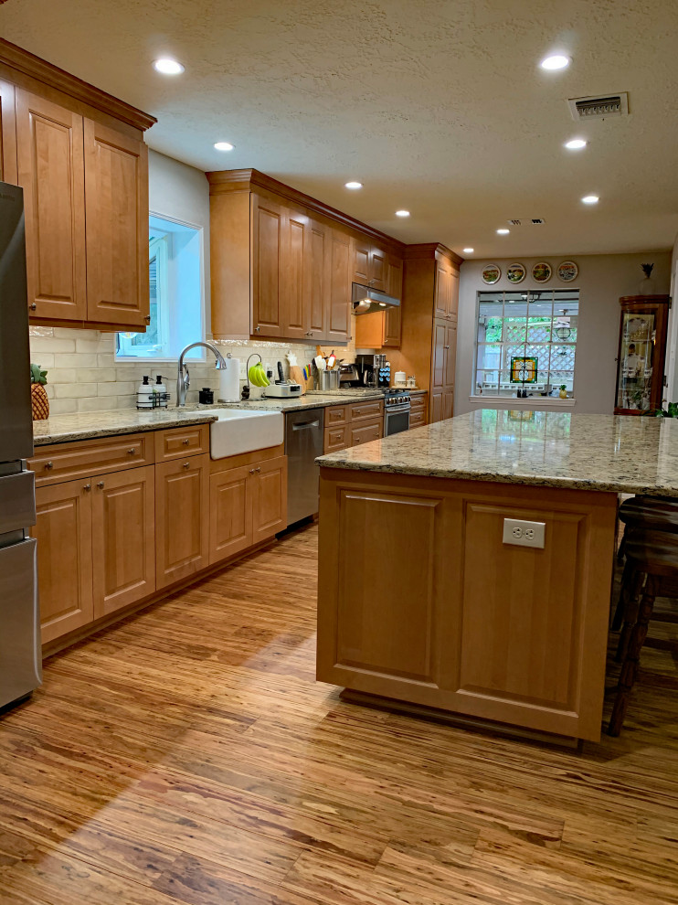 This is an example of a large traditional single-wall open plan kitchen in Houston with a farmhouse sink, raised-panel cabinets, light wood cabinets, solid surface benchtops, beige splashback, subway tile splashback, stainless steel appliances, vinyl floors, with island, multi-coloured floor and brown benchtop.