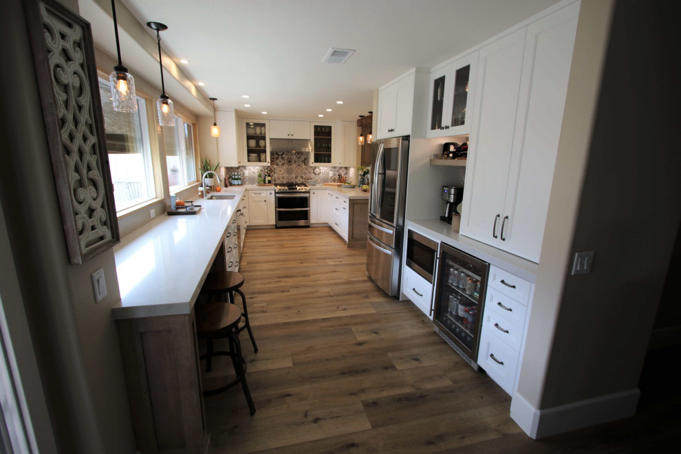 Photo of a large transitional u-shaped kitchen pantry in Orange County with a farmhouse sink, shaker cabinets, white cabinets, granite benchtops, brown splashback, ceramic splashback, stainless steel appliances, light hardwood floors, with island, brown floor, white benchtop and vaulted.