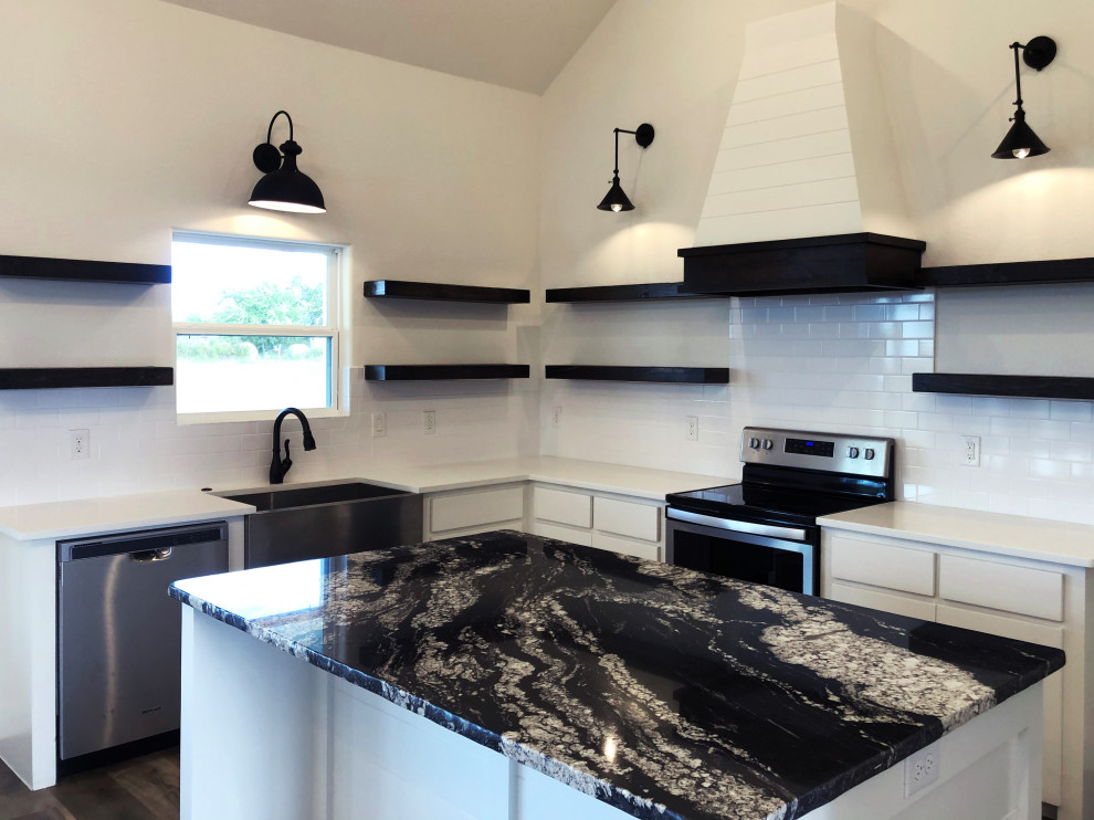 Medium sized country l-shaped open plan kitchen in Dallas with a belfast sink, shaker cabinets, white cabinets, granite worktops, white splashback, metro tiled splashback, stainless steel appliances, an island, brown floors and black worktops.