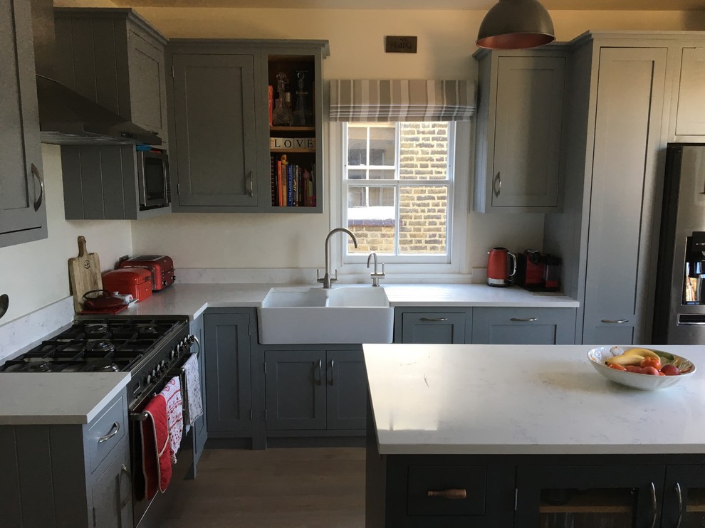 Inspiration for a medium sized traditional l-shaped kitchen/diner in London with a belfast sink, shaker cabinets, grey cabinets, quartz worktops, white splashback, glass sheet splashback, an island, brown floors, stainless steel appliances, light hardwood flooring and white worktops.