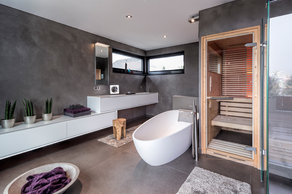 Mid-sized contemporary bathroom in Dusseldorf with flat-panel cabinets, white cabinets, a freestanding tub, a curbless shower, a wall-mount toilet, gray tile, grey walls, with a sauna, grey floor, concrete floors, an undermount sink and a hinged shower door.