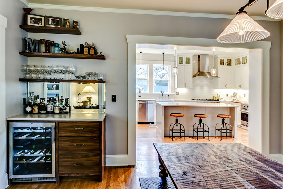 Mid-sized industrial l-shaped eat-in kitchen in Portland with a farmhouse sink, shaker cabinets, white cabinets, quartz benchtops, white splashback, ceramic splashback, stainless steel appliances, medium hardwood floors, with island and brown floor.