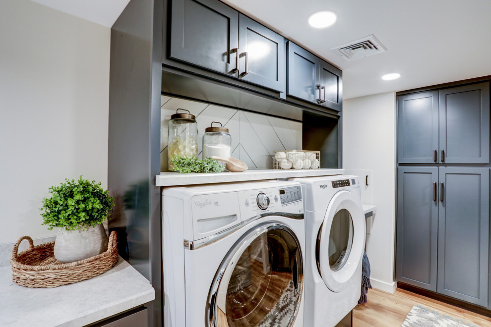 Klassisk inredning av en stor grå l-formad grått tvättstuga enbart för tvätt, med en nedsänkt diskho, luckor med infälld panel, blå skåp, laminatbänkskiva, vita väggar, vinylgolv, en tvättmaskin och torktumlare bredvid varandra och brunt golv