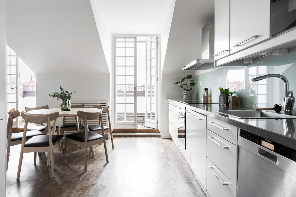 Inspiration for a mid-sized scandinavian single-wall open plan kitchen in Stockholm with medium hardwood floors, brown floor, a drop-in sink, flat-panel cabinets, white cabinets, blue splashback, glass sheet splashback and stainless steel appliances.
