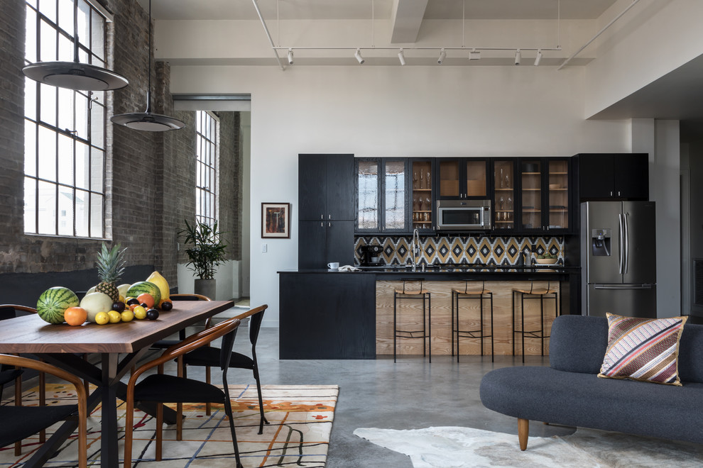 This is an example of an expansive industrial single-wall open plan kitchen in New Orleans with flat-panel cabinets, black cabinets, multi-coloured splashback, ceramic splashback, stainless steel appliances, concrete floors, with island, grey floor and black benchtop.