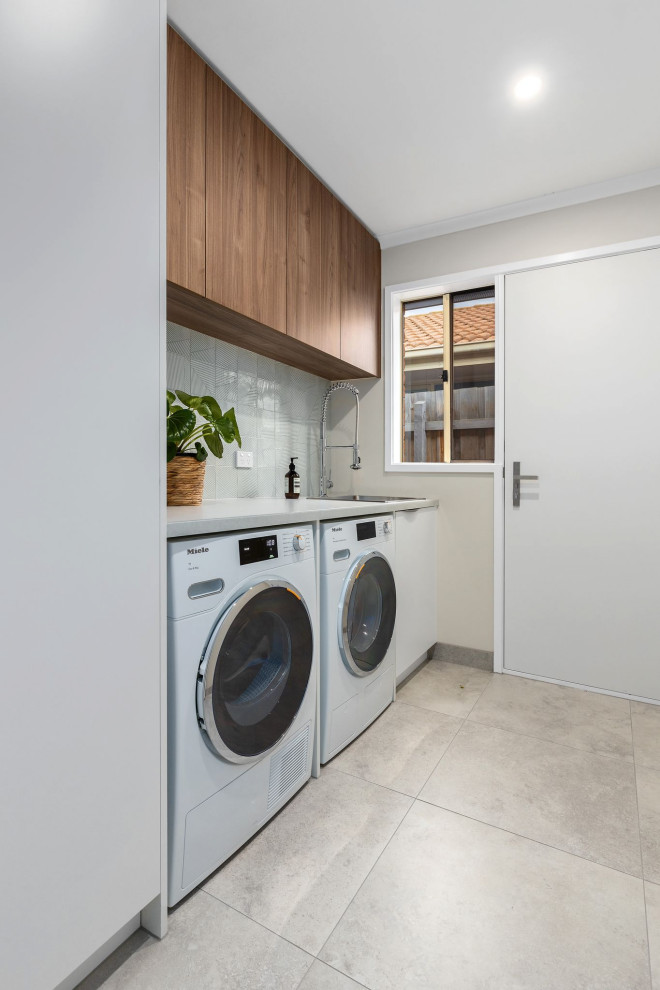 Photo of a mid-sized contemporary single-wall dedicated laundry room in Melbourne with flat-panel cabinets, light wood cabinets, grey walls, porcelain floors, grey floor, a drop-in sink, quartz benchtops, grey splashback, porcelain splashback, a side-by-side washer and dryer and white benchtop.