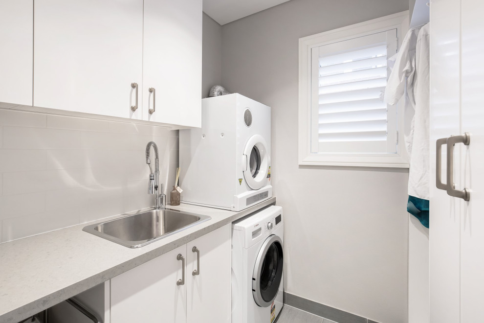 This is an example of a mid-sized transitional single-wall dedicated laundry room in Sydney with a drop-in sink, flat-panel cabinets, white cabinets, laminate benchtops, white walls, porcelain floors, a stacked washer and dryer and grey floor.