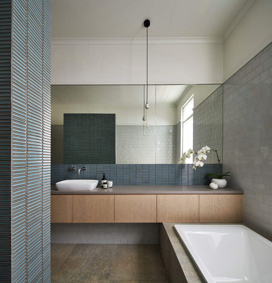 This is an example of a contemporary bathroom in Sydney with medium wood cabinets, a drop-in tub, blue tile, gray tile, white walls, a vessel sink, grey floor and grey benchtops.