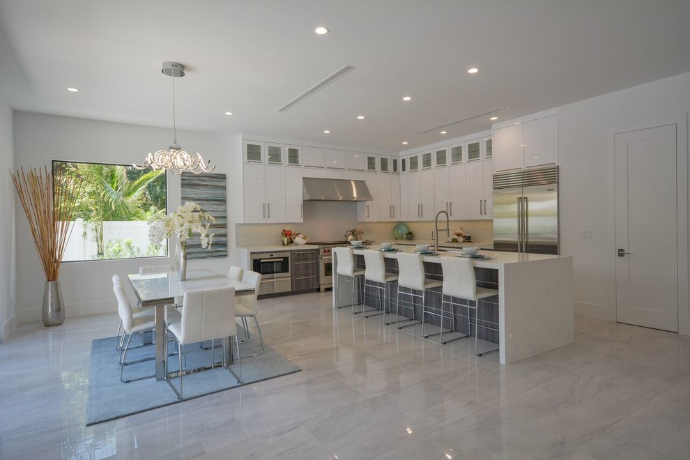 This is an example of a large contemporary l-shaped open plan kitchen in Miami with a farmhouse sink, flat-panel cabinets, grey cabinets, solid surface benchtops, white splashback, glass tile splashback, stainless steel appliances, porcelain floors, with island and white floor.