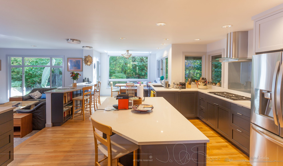 This is an example of a large transitional separate kitchen in Seattle with a double-bowl sink, shaker cabinets, grey cabinets, quartz benchtops, grey splashback, glass sheet splashback, light hardwood floors, with island and stainless steel appliances.