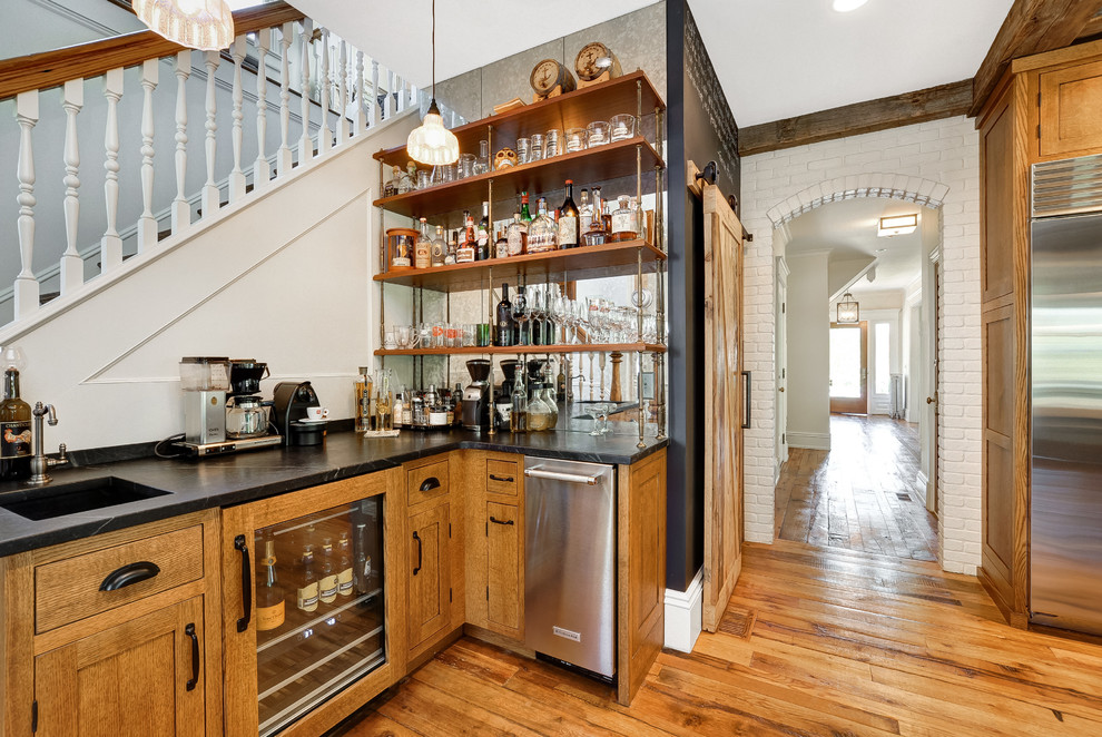 This is an example of a traditional l-shaped wet bar in New York with an integrated sink, shaker cabinets, medium wood cabinets, medium hardwood floors, brown floor and black benchtop.