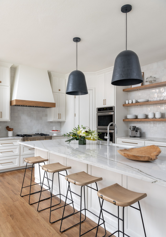 Inspiration for a mid-sized transitional l-shaped open plan kitchen in Dallas with a farmhouse sink, recessed-panel cabinets, white cabinets, quartzite benchtops, white splashback, ceramic splashback, stainless steel appliances, medium hardwood floors, with island and white benchtop.