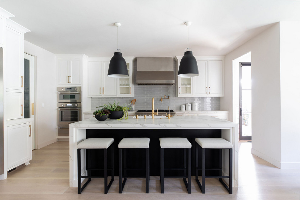 Mid-sized transitional l-shaped light wood floor eat-in kitchen photo in Sacramento with a farmhouse sink, shaker cabinets, white cabinets, gray backsplash, ceramic backsplash, stainless steel appliances, an island and white countertops