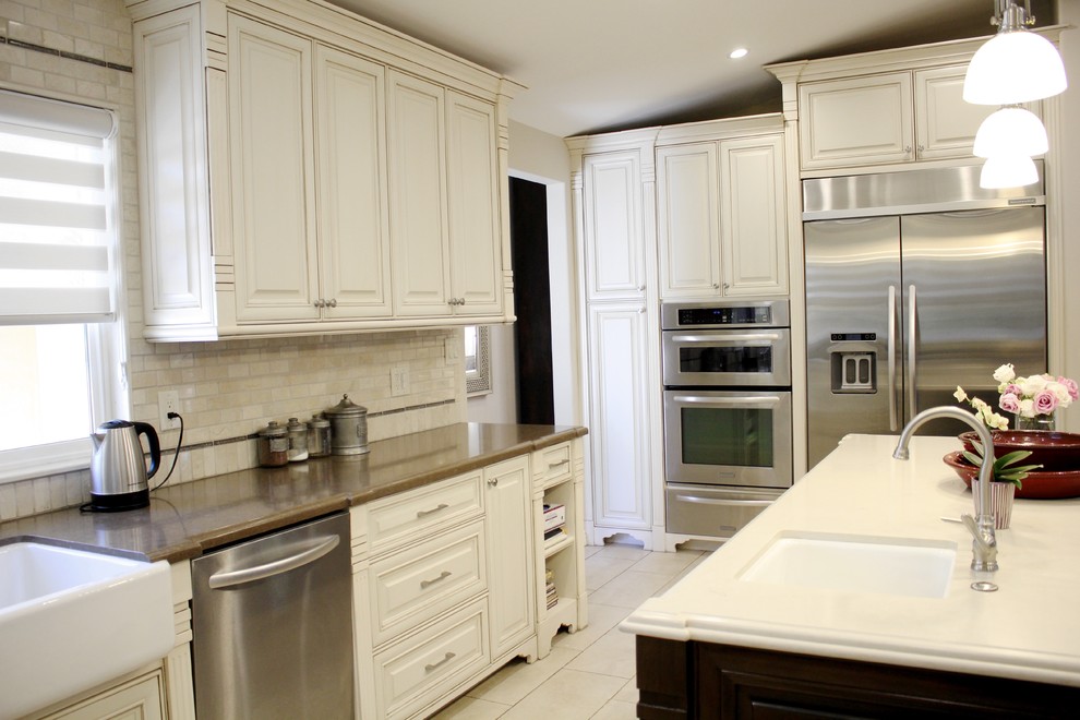Mid-sized traditional l-shaped eat-in kitchen in Los Angeles with a farmhouse sink, shaker cabinets, beige cabinets, quartz benchtops, beige splashback, travertine splashback, stainless steel appliances, marble floors, with island, white floor and beige benchtop.