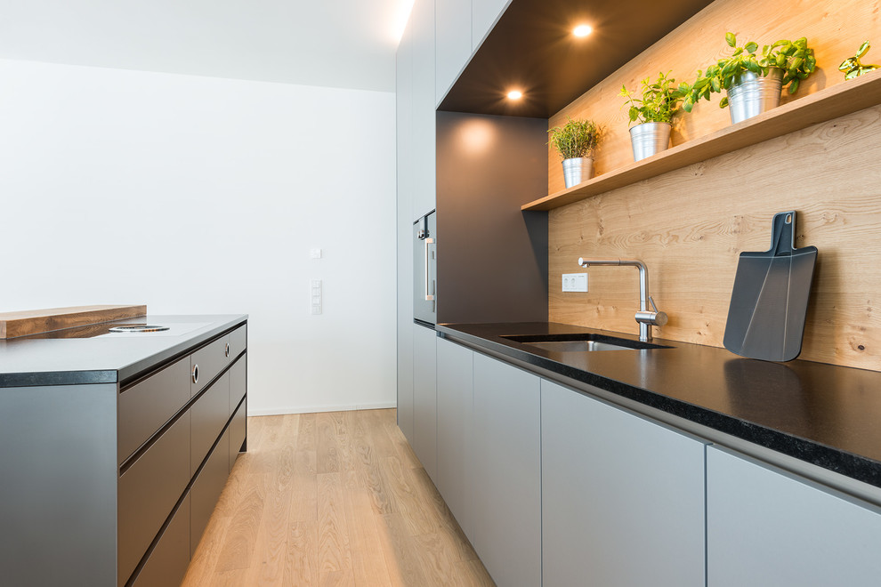 Küche mit grauen Schränken, Rückwand aus Holz, schwarzen Elektrogeräten, Kücheninsel, braunem Boden, schwarzer Arbeitsplatte, Unterbauwaschbecken, flächenbündigen Schrankfronten, Granit-Arbeitsplatte, Küchenrückwand in Braun und braunem Holzboden in Munich