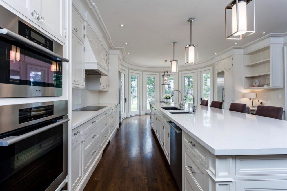 Inspiration for a large traditional galley open plan kitchen in Toronto with an undermount sink, recessed-panel cabinets, white cabinets, quartz benchtops, stainless steel appliances, dark hardwood floors, with island, brown floor and white benchtop.