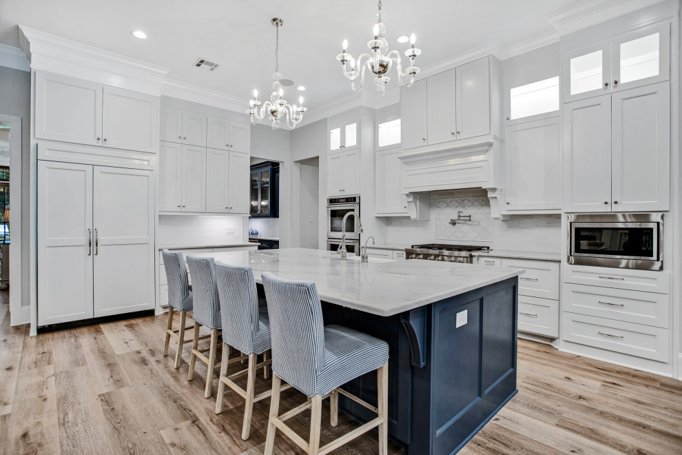 Design ideas for a large l-shaped open plan kitchen in Houston with a belfast sink, recessed-panel cabinets, white cabinets, white splashback, metro tiled splashback, stainless steel appliances, medium hardwood flooring, an island, brown floors and grey worktops.