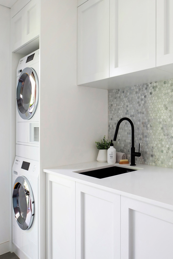 Design ideas for a mid-sized beach style galley dedicated laundry room in Sydney with an undermount sink, shaker cabinets, white cabinets, quartz benchtops, grey splashback, mosaic tile splashback, white walls, porcelain floors, a stacked washer and dryer, grey floor, white benchtop, coffered and panelled walls.