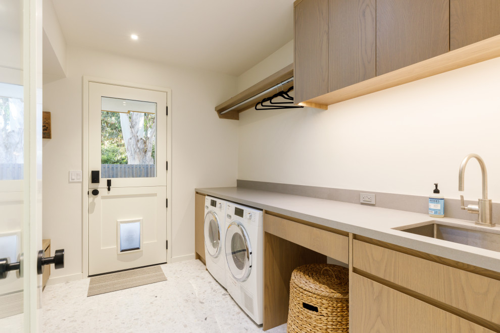 Idéer för mellanstora funkis parallella grått tvättstugor enbart för tvätt, med en nedsänkt diskho, släta luckor, bruna skåp, vita väggar, ljust trägolv, en tvättmaskin och torktumlare bredvid varandra och brunt golv