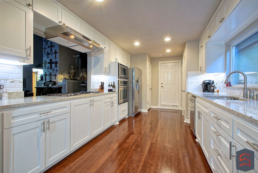 This is an example of a mid-sized transitional galley separate kitchen in Dallas with white cabinets, granite benchtops, white splashback, stainless steel appliances, a double-bowl sink, shaker cabinets, subway tile splashback and dark hardwood floors.