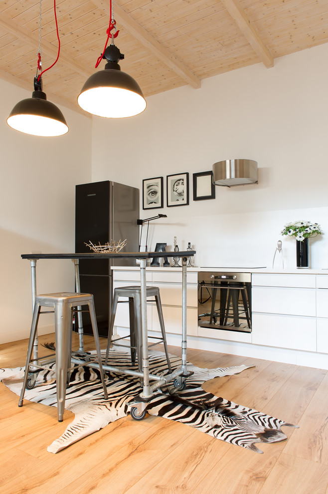 This is an example of a mid-sized industrial single-wall eat-in kitchen in Dusseldorf with flat-panel cabinets, white cabinets, medium hardwood floors, white splashback, no island and black appliances.