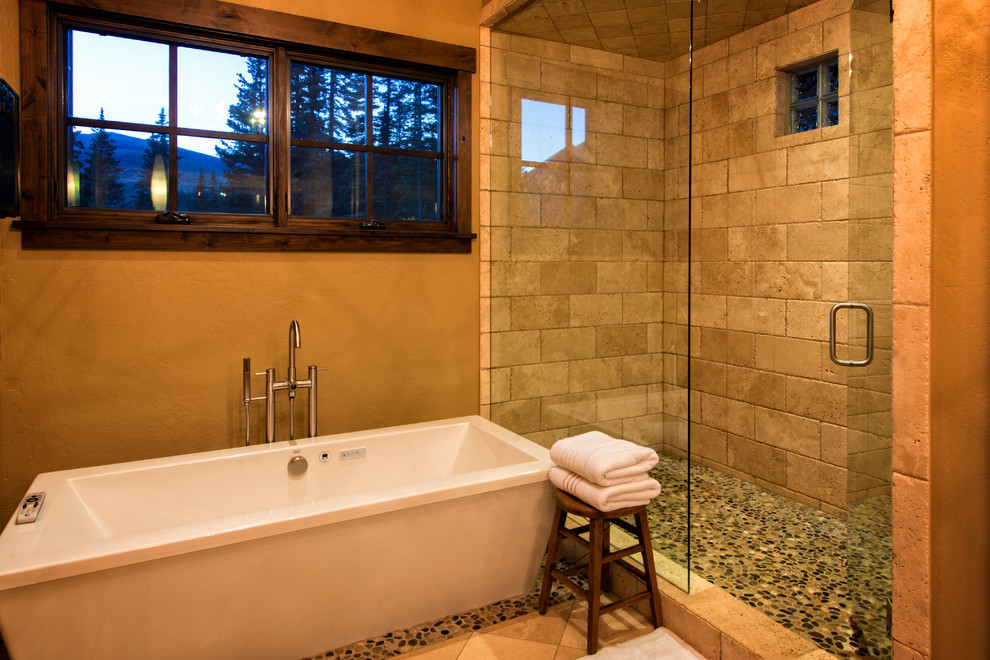 Photo of a large country master bathroom in Denver with a freestanding tub, an alcove shower, beige tile, stone tile, brown walls, pebble tile floors and brown floor.