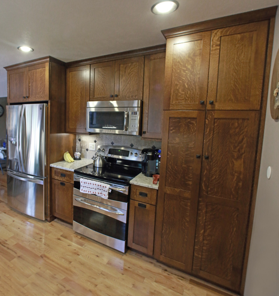 Photo of a small country l-shaped eat-in kitchen in Other with a farmhouse sink, shaker cabinets, brown cabinets, granite benchtops, beige splashback, ceramic splashback, stainless steel appliances, light hardwood floors, a peninsula and yellow floor.