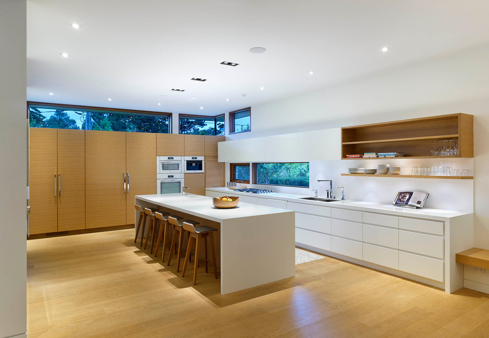 Design ideas for a large modern l-shaped open plan kitchen in Toronto with an undermount sink, flat-panel cabinets, white cabinets, solid surface benchtops, white splashback, white appliances, light hardwood floors and with island.