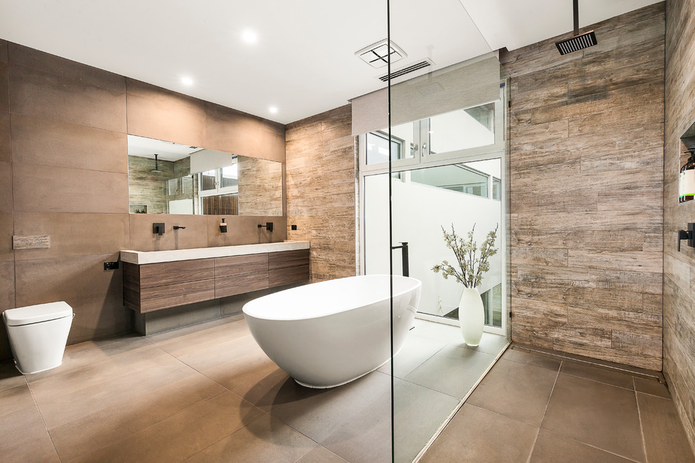 This is an example of a large contemporary master bathroom in Melbourne with a one-piece toilet, gray tile, brown tile, concrete benchtops, an open shower, flat-panel cabinets, brown cabinets, a freestanding tub, brown walls, brown floor, a corner shower, a wall-mount sink and grey benchtops.