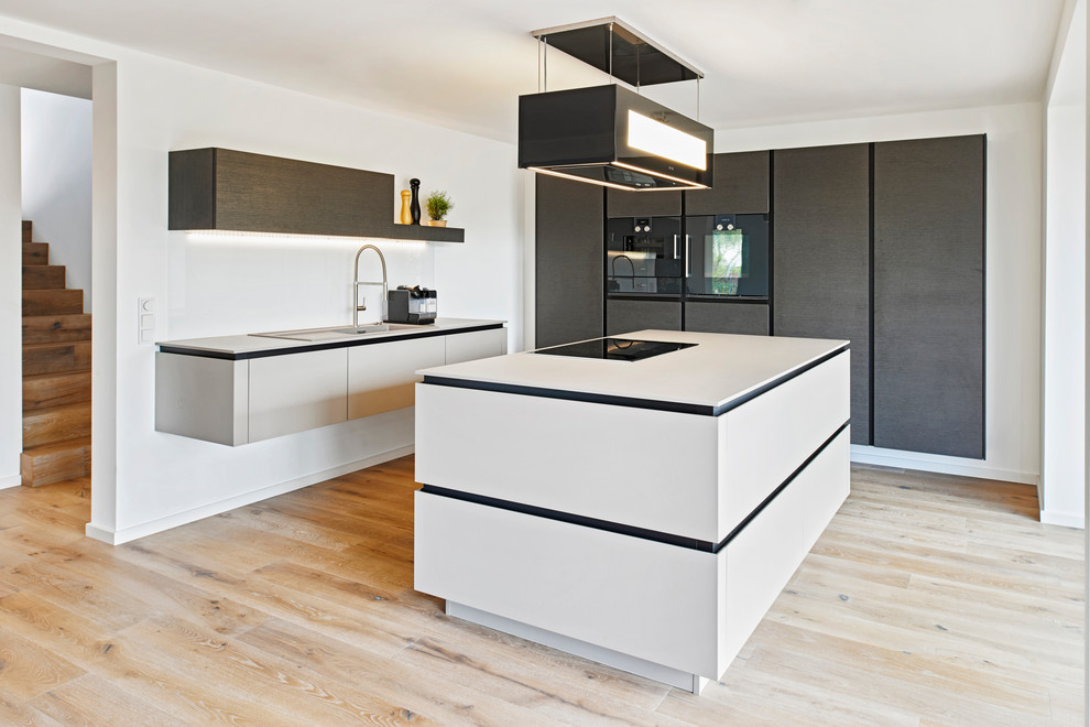 This is an example of a mid-sized contemporary l-shaped open plan kitchen in Stuttgart with a drop-in sink, flat-panel cabinets, black cabinets, white splashback, light hardwood floors, with island and black appliances.