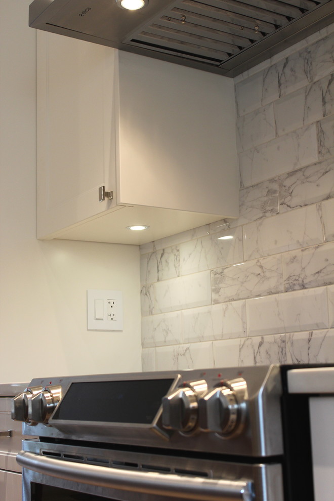 Large transitional l-shaped light wood floor home design photo with an undermount sink, shaker cabinets, white cabinets, quartz countertops, white backsplash, ceramic backsplash, stainless steel appliances and an island